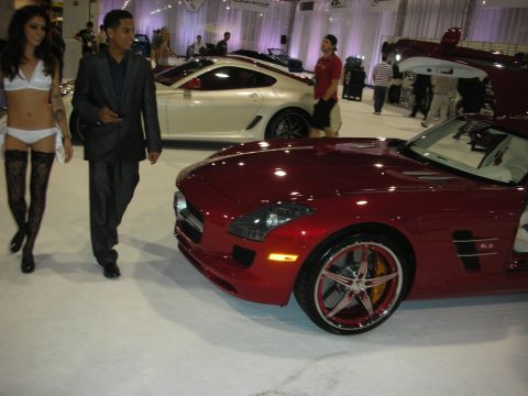 1998 Ford Crown Victoria - red Mercedes-Benz SLS AMG with 6.3 fender badge and chrome wheel on a show floor as a couple and other attendees walk past.