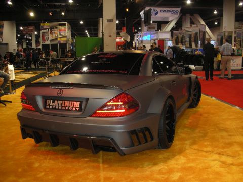 1998 Ford Crown Victoria - matte-gray widebody Mercedes coupe with Platinum Motorsport windshield and trunk graphics on black wheels at an indoor expo hall.