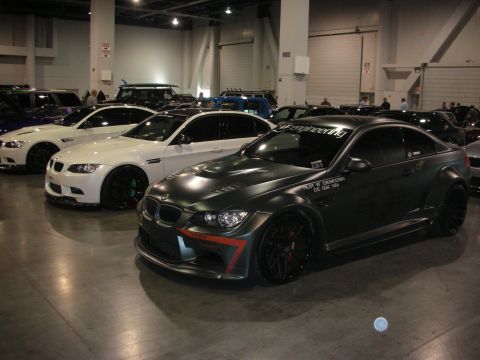 1998 Ford Crown Victoria - row of BMW 3 Series coupes on a show floor, led by a matte dark-green widebody car beside a white one with custom wheels.
