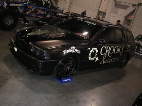 1998 Ford Crown Victoria - matte-black BMW 5 Series wagon wrapped with Crooks & Castles and Falken graphics, on black wheels resting on tire-brand display stands.