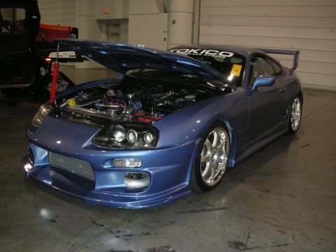 1998 Ford Crown Victoria - blue Toyota Supra with body kit and rear wing, hood open to reveal the engine bay, parked on a concrete show floor.