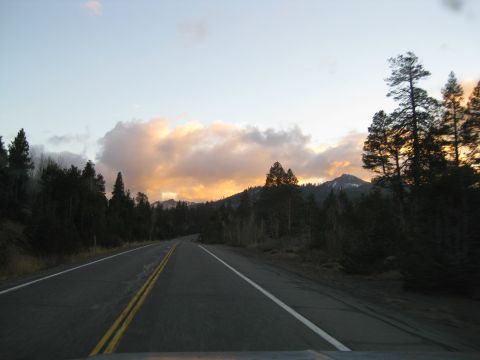 1986 Ford Bronco - A road winds through a forest with mountains in the distance under a colorful sunset sky.