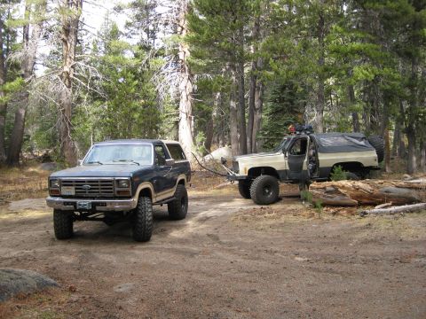 1986 Ford Bronco - A Ford Bronco is towed by a Chevrolet K5 Blazer on a dirt road surrounded by trees.