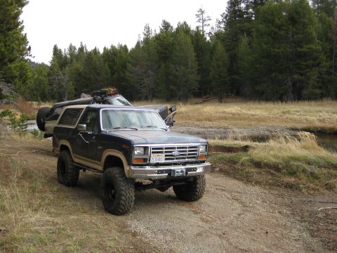 1986 Ford Bronco - A dark blue Ford Bronco is parked on a dirt road, with another vehicle behind it in a wooded area.