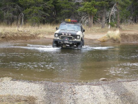 1986 Ford Bronco - This modified off-road vehicle navigates through a stream in a wooded area.