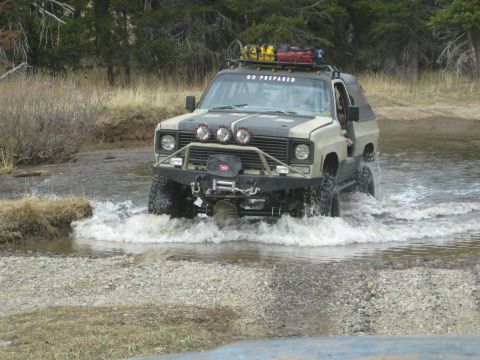 1986 Ford Bronco - Modified SUV navigates a shallow stream, equipped for adventure with extra fuel cans and lights.