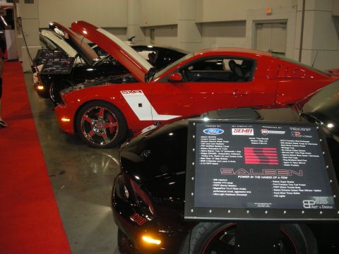 1998 Ford Crown Victoria - red and white Saleen Mustang with hood open on a show floor, displayed next to a black Saleen spec board and a black car.