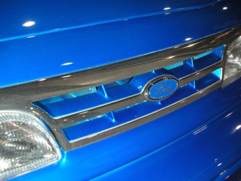 1998 Ford Crown Victoria - Close-up of a glossy blue vehicle's chrome-trimmed grille centered on a Ford oval badge beside the headlight.