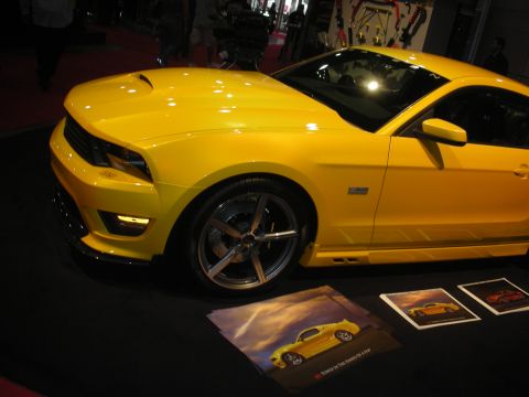 1998 Ford Crown Victoria - yellow modern muscle coupe shown in profile with staggered wheels and promo posters on the floor at an indoor show.
