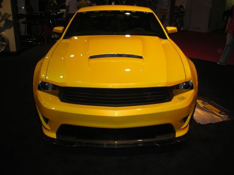 1998 Ford Crown Victoria - Head-on view of a yellow modern muscle car with a center hood scoop and blacked-out grille on a show display stand.