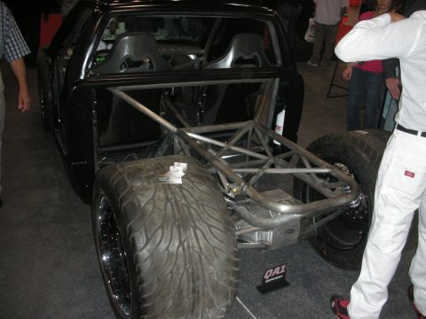 1998 Ford Crown Victoria - stripped tube-frame race car showing its exposed rear suspension, tube chassis and wide rear tire at a show booth.