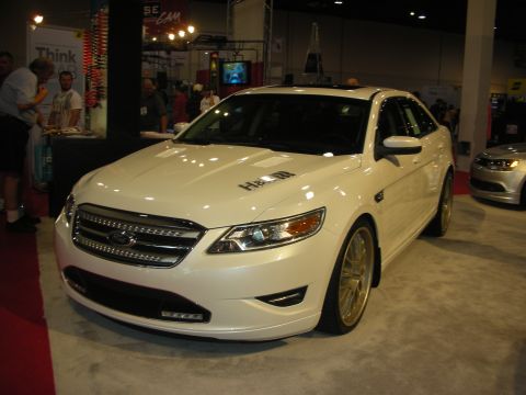1998 Ford Crown Victoria - white Ford full-size sedan lowered on gold mesh wheels displayed at an indoor automotive show booth.