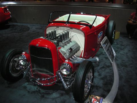 1998 Ford Crown Victoria - red highboy hot rod roadster with white interior and a V8 wearing tall chrome velocity-stack intakes, displayed on show carpet.