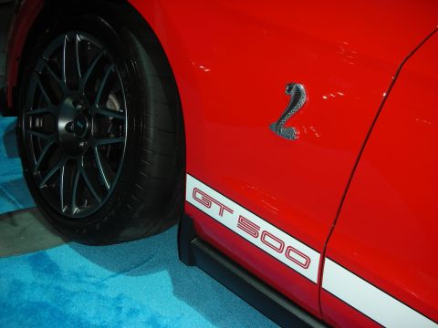 1998 Ford Crown Victoria - red car's rocker shows a white GT 500 stripe and a chrome Shelby cobra emblem above a black multi-spoke wheel.