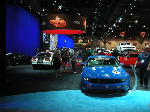 1998 Ford Crown Victoria - busy indoor auto show floor with a numbered blue race coupe in the foreground and attendees walking among other display cars.