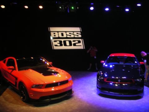 1998 Ford Crown Victoria - orange coupe and a dark blue coupe staged under spotlights below an illuminated BOSS 302 sign.