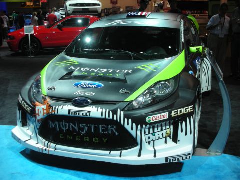 1998 Ford Crown Victoria - Ford Fiesta rally car in black, white and green Monster Energy livery with sponsor decals displayed on a blue stand.