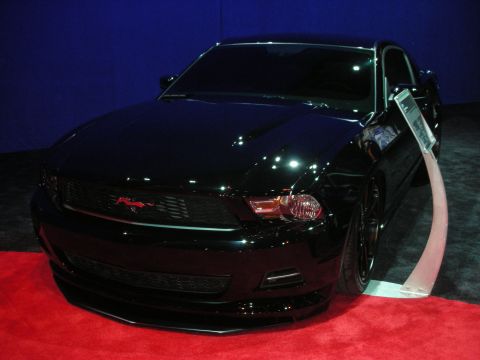 1998 Ford Crown Victoria - glossy black Ford Mustang shown front three-quarter on a red carpet with a blue backdrop and an info tag hanging at the door.