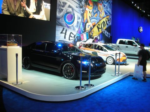 1998 Ford Crown Victoria - black lowered Ford sedan on a white display stand in front of a colorful graffiti-style mural, with a pickup nearby.