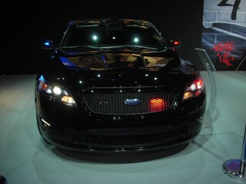 1998 Ford Crown Victoria - Head-on view of a black Ford sedan with a chrome-trimmed mesh grille and projector lights on an illuminated white display stand.