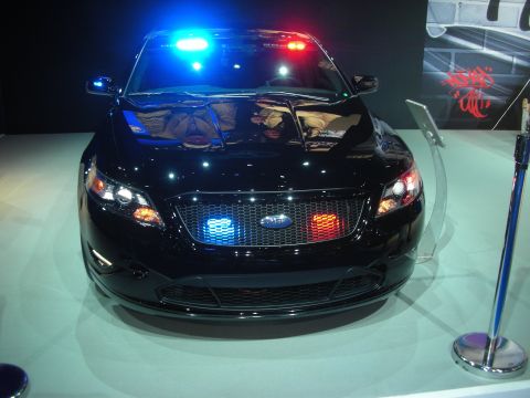 1998 Ford Crown Victoria - black Ford sedan configured as a police car with a roof-mounted light bar and red and blue grille lights on a show stand.