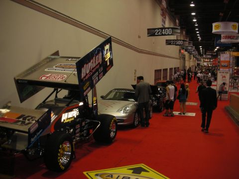 1998 Ford Crown Victoria - red-carpeted trade show aisle lined with displays, including a sprint car with a large wing and a silver sports car, as attendees walk by.