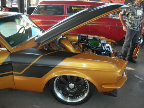 1998 Ford Crown Victoria - gold and black custom muscle car with its hood open revealing the engine, sitting on black multi-spoke wheels at an outdoor show.