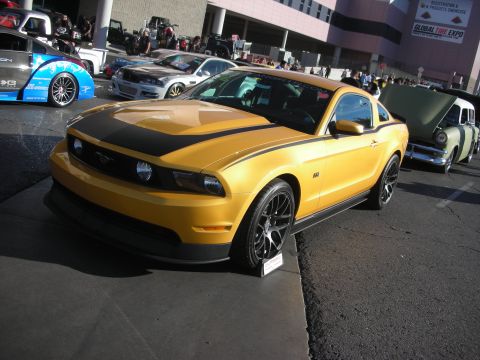 1998 Ford Crown Victoria - gold Ford Mustang coupe with a black hood stripe and black wheels parked outdoors among other vehicles at an auto event.