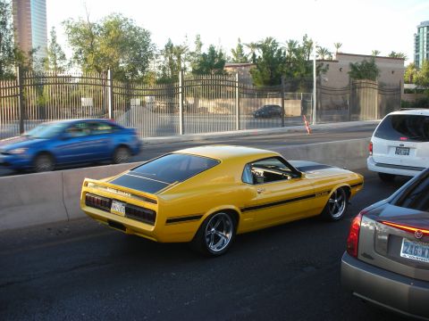 1998 Ford Crown Victoria - yellow fastback muscle car with black side stripes and chrome wheels parked along a fenced city street in traffic.