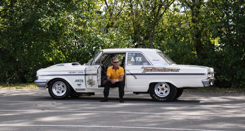 White Thunderbolt drag car in side view with door open and man seated inside, parked before trees