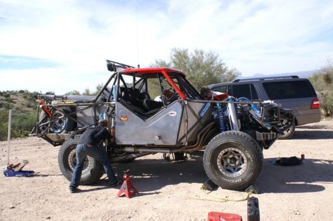1990 Ford Bronco - tube-frame off-road race car with bypass shocks sits on jack stands in the desert as a mechanic works near the wheels.