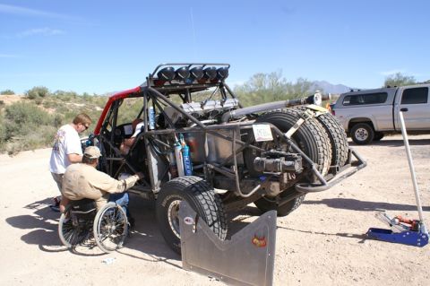 1990 Ford Bronco - Rear view of a single-seat tube-frame off-road racer with roof light bar and spare tire while people work alongside it in the desert.