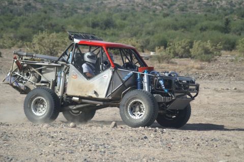 1990 Ford Bronco - tube-frame off-road race car with a red roof section drives over rocky desert terrain, raising dust.