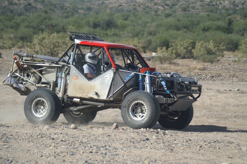 Tube-chassis off-road race car with red roof driving through desert kicking up dust