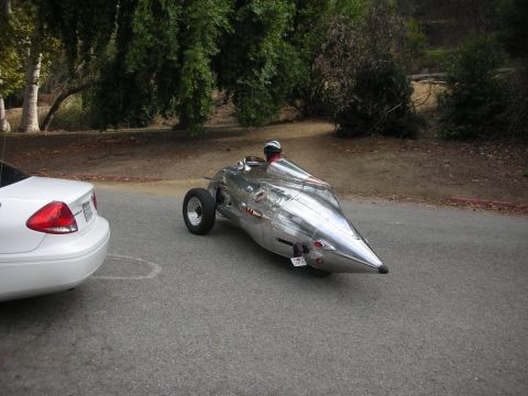 2004 Ford Crown Victoria - A custom streamliner race car is parked on a paved road next to a white sedan.