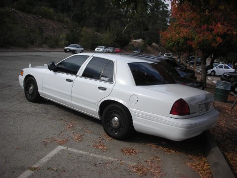 2004 Ford Crown Victoria - A white 2004 Ford Crown Victoria is parked in a lot with other vehicles and trees in the background.