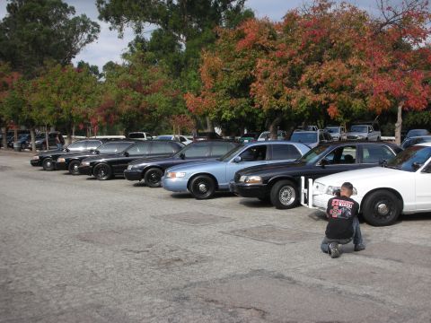 2004 Ford Crown Victoria - A photographer kneels to capture the front of a white police car, with several other similar vehicles parked in a row.