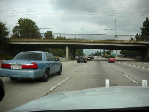 2004 Ford Crown Victoria - A light blue sedan is seen from behind on a highway, driving under a concrete overpass.