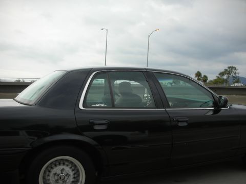 2004 Ford Crown Victoria - A black sedan is seen from the side on a highway under a cloudy sky.