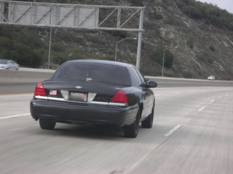 2004 Ford Crown Victoria - A black 2004 Ford Crown Victoria is seen from behind on a multi-lane highway with a hillside in the background.