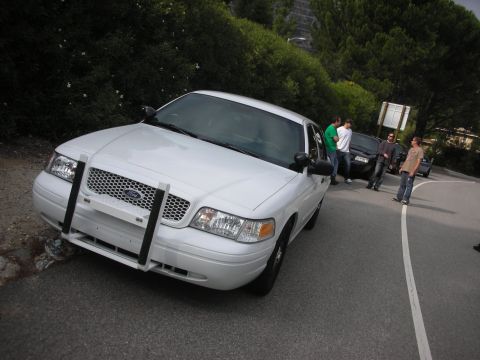 2004 Ford Crown Victoria - A group of people gather around a white Ford Crown Victoria, possibly a police car, parked on the side of a winding road.