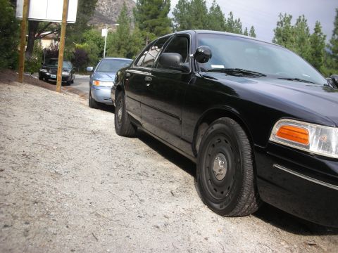 2004 Ford Crown Victoria - Two police interceptor sedans, a black Crown Victoria and a light blue model, are parked on a gravel shoulder.