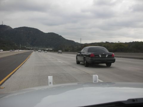 2004 Ford Crown Victoria - A black 2004 Ford Crown Victoria travels down a multi-lane highway under an overcast sky.
