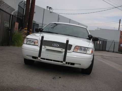 2004 Ford Crown Victoria - A white Ford Crown Victoria police car is parked on an asphalt surface in front of a fence and buildings.