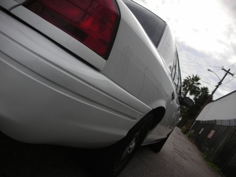 2004 Ford Crown Victoria - The rear of a white sedan is shown from a low angle in a narrow alleyway with a cloudy sky above.