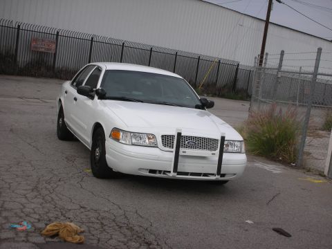 2004 Ford Crown Victoria - A white 2004 Ford Crown Victoria police car is parked on a cracked asphalt surface in front of a fence.