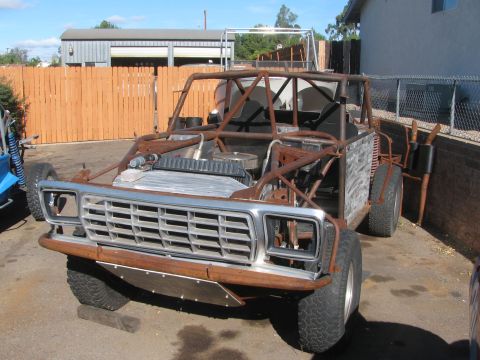 1990 Ford Bronco - tube-frame off-road buggy fitted with a chrome truck grille sits in mid-build on a paved driveway by a wood fence.