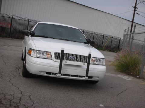 2004 Ford Crown Victoria - A white 2004 Ford Crown Victoria is parked on a cracked asphalt surface, facing forward with a push bar on its grille.