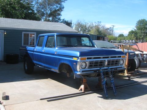 1990 Ford Bronco - blue Ford crew-cab pickup with FORD lettering on the hood sits raised on jack stands over blue coilovers in a driveway.