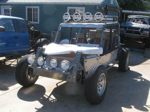 1990 Ford Bronco - bare-aluminum off-road buggy with four Hella roof lights and round front lights parked among other vehicles on a driveway.